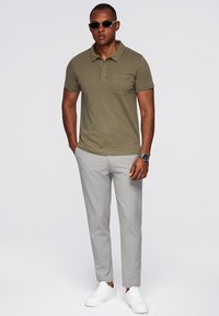Polo shirt verde oliva con taschino, abbinato a pantaloni grigi chiari slim-fit e sneakers bianche. Il modello indossa occhiali da sole.