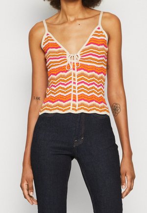 Frau, die ein ärmelloses Strickoberteil mit orangefarbenen, pinken und weißen Zickzackstreifen und dunkle hoch taillierte Jeans trägt.