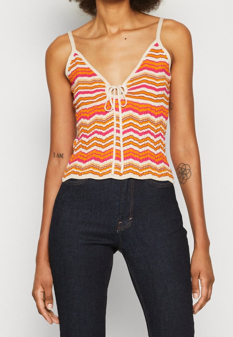 Femme portant un haut en tricot sans manches avec des rayures zigzag orange, rose et blanche et un jean taille haute foncé.