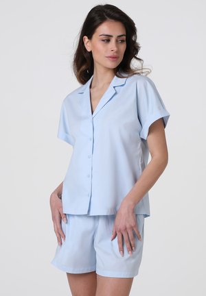 Donna che indossa una camicia azzurra a maniche corte con bottoni e pantaloncini coordinati, in piedi con le mani appoggiate vicino alle cosce, guarda leggermente di lato.