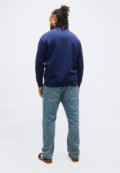 Άνδρας με ντραντλοκς που φοράει μπλε σκούρο φούτερ, ανοιχτό μπλε τζιν και μαύρα-λευκά παπούτσια, στέκεται μπροστά σε απλό λευκό φόντο.