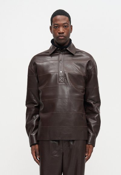 Férfi sötétbarna bőrlong sleeve pólót és hozzá illő bőr nadrágot visel, egy sima világos háttér előtt állva.