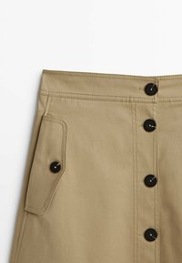 Jupe beige avec boutons noirs sur le devant et une poche latérale avec rabat et fermeture par bouton, coutures détaillées visibles.