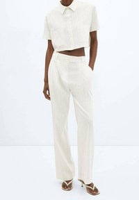 Personne à la peau foncée portant une chemise courte à manches courtes cropped crème à fines rayures et un pantalon large taille haute assorti, avec des sandales à talons beiges.