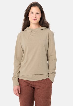 Vrouw met lang bruin haar, gekleed in een beige hoodie en roestkleurige broek, staand tegen een effen lichte achtergrond.