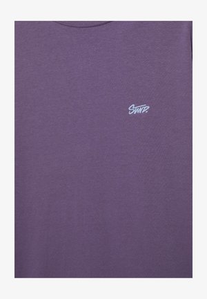 Lila T-Shirt mit Rundhalsausschnitt und kleinem weißem gesticktem Schriftzug "SWTD." auf der linken Brustseite.