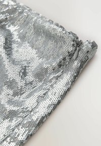 Nahaufnahme von silbernem Paillettenstoff mit einem geschlossenen Reißverschlussanhänger vor einem schlichten hellen Hintergrund.