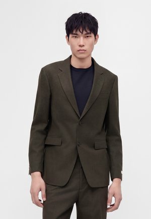 Jeune homme portant une veste et un pantalon de costume vert olive foncé avec une chemise noire en dessous, debout devant un fond blanc uni.