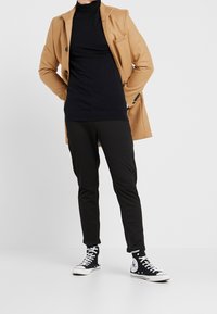 Person in einem kamelbraunen Mantel über einem schwarzen Rollkragenpullover, schwarzen Hosen und schwarzen Converse High-Top-Sneakern steht vor einem weißen Hintergrund.