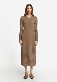 Långärmad, brun stickad klänning med texturerad öppenvävsdesign, en figurnära midja och utsvängd nederkant, kombinerad med bruna platta sandaler.