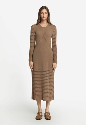 Långärmad, brun stickad klänning med texturerad öppenvävsdesign, en figurnära midja och utsvängd nederkant, kombinerad med bruna platta sandaler.