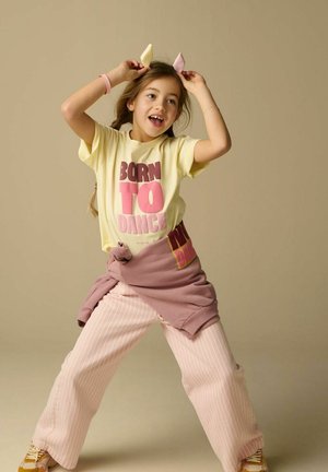 Jong meisje danst vrolijk, gekleed in een T-shirt met de tekst "Born to Dance", gestreepte broek, geknoopte trui en een speels hoofdbandje met kattenoren.