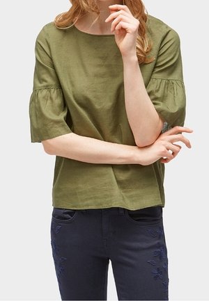 Kvinde iført olivengrøn kortærmet bluse med puffede ærmer og marineblå bukser med blomsterbroderi på lårene.