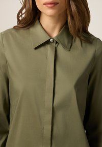 Camicia verde oliva con bottoni, realizzata in tessuto liscio, con colletto a punta e un'unica bottone decorativo al collo.