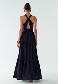 Jūras zila maksi kleita ar teksturētu apdari, ar mezglotu detaļu mugurā un daudzslāņu svārku dizainu. Gludā auduma drapējums ir elegants un plūstošs.