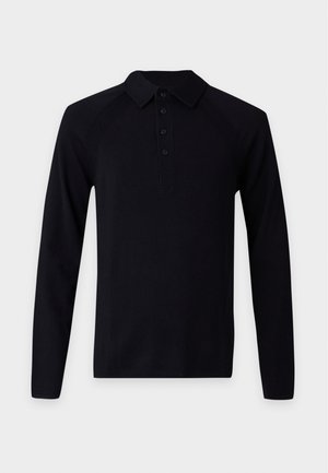 Man met een donkere huid, gekleed in een zwart poloshirt met lange mouwen, lichtblauwe wijde jeans en tan schoenen, staand tegen een effen lichte achtergrond.