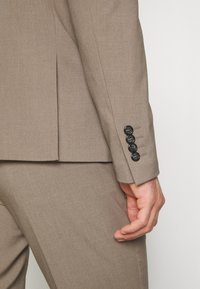 Brown tailored suit jacket featuring a textured fabric, a single vent, and four black buttons on the cuff. Visible hand in a relaxed position.