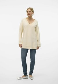 Pull oversized crème en v avec un col et des poignets côtelés, associé à un jean bleu slim et des ballerines argentées, sur fond blanc.
