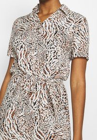 Donna che indossa un vestito corto con maniche corte, con bottoni e vita annodata, in un motivo astratto di stampa animale beige, nero e bianco.