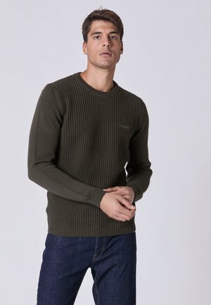Maglione a coste di un verde scuro con collo a giro e maniche lunghe, abbinato a jeans in denim scuro. Il dettaglio del tessuto testurizzato è visibile.