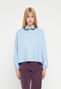 Lyseblå skjorte med knapper, gr�øn broderet krave, lange ærmer og kort snit. Stoffet ser glat og blødt ud.