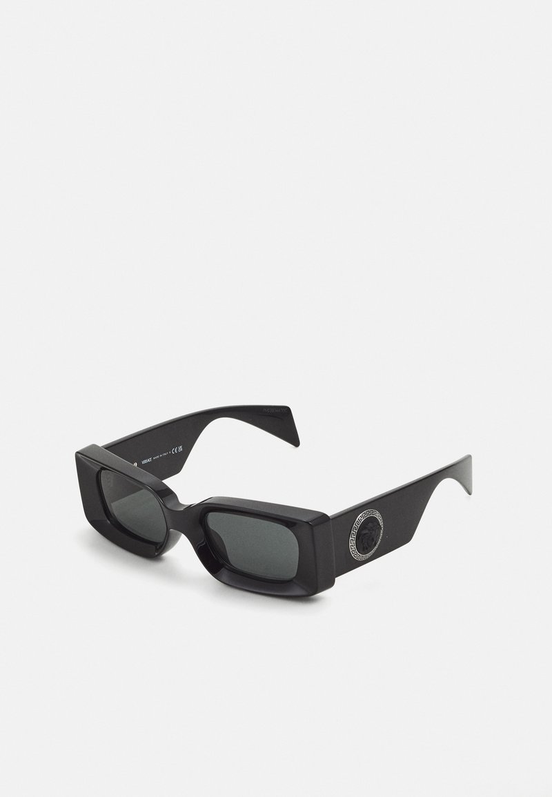 Lunettes de soleil noires avec des montures rectangulaires épaisses, des verres sombres, des branches courbées et un accent géométrique rond sur le côté. Texture en plastique lisse.