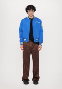 Chaqueta bomber azul con rayas amarillas, camiseta negra, pantalones de satén marrón y zapatos negros, con detalles bordados y grandes gafas.