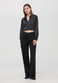 Camisetta nera a pois con maniche lunghe, abbinata a jeans neri a vita alta svasati, che mostrano un design aderente e una superficie morbida.