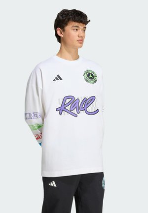 Jeune homme portant une chemise blanche à manches longues avec le texte "Race" et des graphismes colorés sur les manches, associée à un pantalon noir avec des écussons de logo.