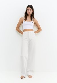 Femme aux longs cheveux bruns porte un crop top blanc et un pantalon blanc taille haute à jambes larges, debout pieds nus contre un mur blanc.