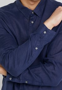 Persona che indossa una camicia a maniche lunghe blu navy con colletto, braccia incrociate sul petto, e tonalità di pelle chiara visibile su mano e collo.