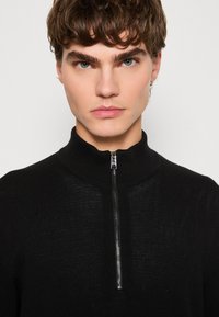 Black knit sweater with a high collar and a half zipper. The texture is subtle, and the design is minimalist, emphasizing simplicity.