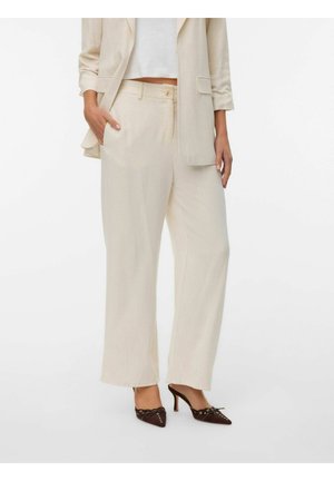 Žena odjevena u bež široke hlače, odgovarajući sako s podvrnutim rukavima, bijeli skraćeni top i tamno smeđe šiljate štikle.