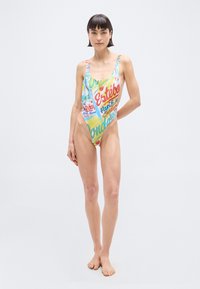 Γυναίκα που στέκεται ξυπόλητη, φορώντας πολύχρωμο ολόσωμο μαγιό με ψηλό κόψιμο και σχέδια κειμένων και γραφικών σε απλό φόντο.