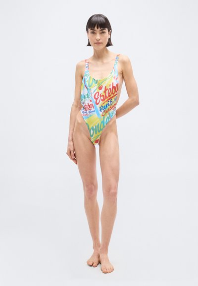 Γυναίκα που στέκεται ξυπόλητη, φορώντας πολύχρωμο ολόσωμο μαγιό με ψηλό κόψιμο και σχέδια κειμένων και γραφικών σε απλό φόντο.