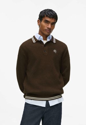 Noor mees kannab pruuni tekstuuriga kootud kampsunit, millel on valged triibulised krae ja mansetid, helesinise särke ja tume teksapükste peal, seistes käed taga.