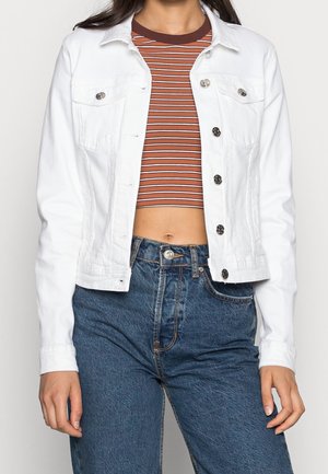 Person trägt eine weiße, geknöpfte Jacke über einem braun-weiß gestreiften Crop-Top und hoch taillierter blauer Jeans, stehend vor neutralem Hintergrund.