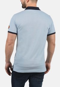 Lys blå polo-shirt med marineblå krage, korte ærmer og brune accenter på skulderen. Lavet af glat stof, tætsiddende snit.