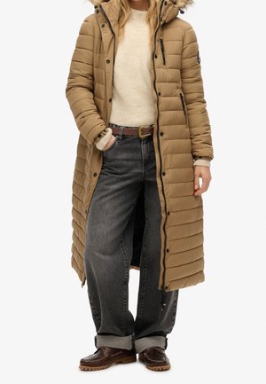 Femme portant un long manteau matelassé beige avec une capuche bordée de fourrure, un pull beige clair, un jean noir retroussé, une ceinture marron et des chaussures marron foncé.