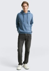 Jauns vīrietis ir ģērbies zilos kapucē, tumši pelēkos džinsos un baltās sporta kurpēs, stāvot ar rokām aiz muguras uz vienkārša fona.