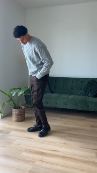 Pilkas kabeliniu mezgimu megztukas, rudos kargo kelnės ir juodi batai. Žalias sofų kampas ir augalas vazonėlyje kambaryje su medinėmis grindimis.