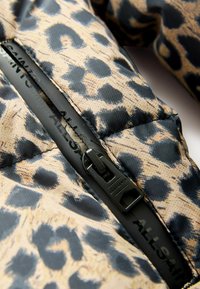 Puff jacket i beige stof med sorte leopardpletter, stor lynlås med branding og quiltet tekstur.