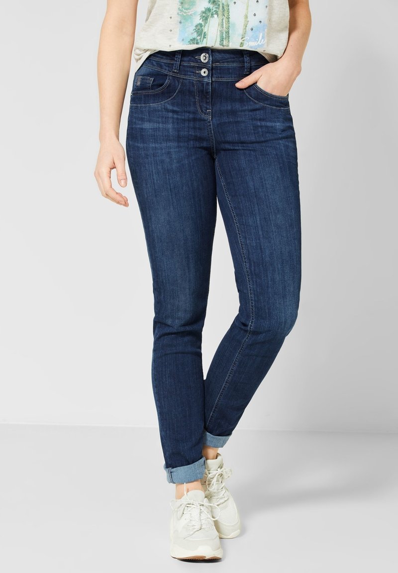 Femme portant un jean skinny bleu foncé retroussé, des baskets blanches et un t-shirt clair avec un imprimé de palmier.