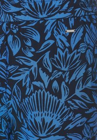 Tissu à motifs floraux bleus avec de grandes fleurs stylisées. Fond bleu marine foncé avec des motifs bleus vibrants et une petite étiquette de marque.