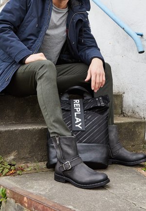 Personne portant des bottes en cuir noires, un pantalon olive, une chemise grise et une veste marine assise sur des marches en béton avec un sac à dos noir marqué "REPLAY".