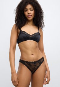 Musta pitsinen bralette kukkakoristeisella reunuksella ja säädettävillä olkaimilla, yhdistettynä läpikuultaviin pitsiseen stringeihin, joissa on kukkaompeleita ja aaltoreunat.