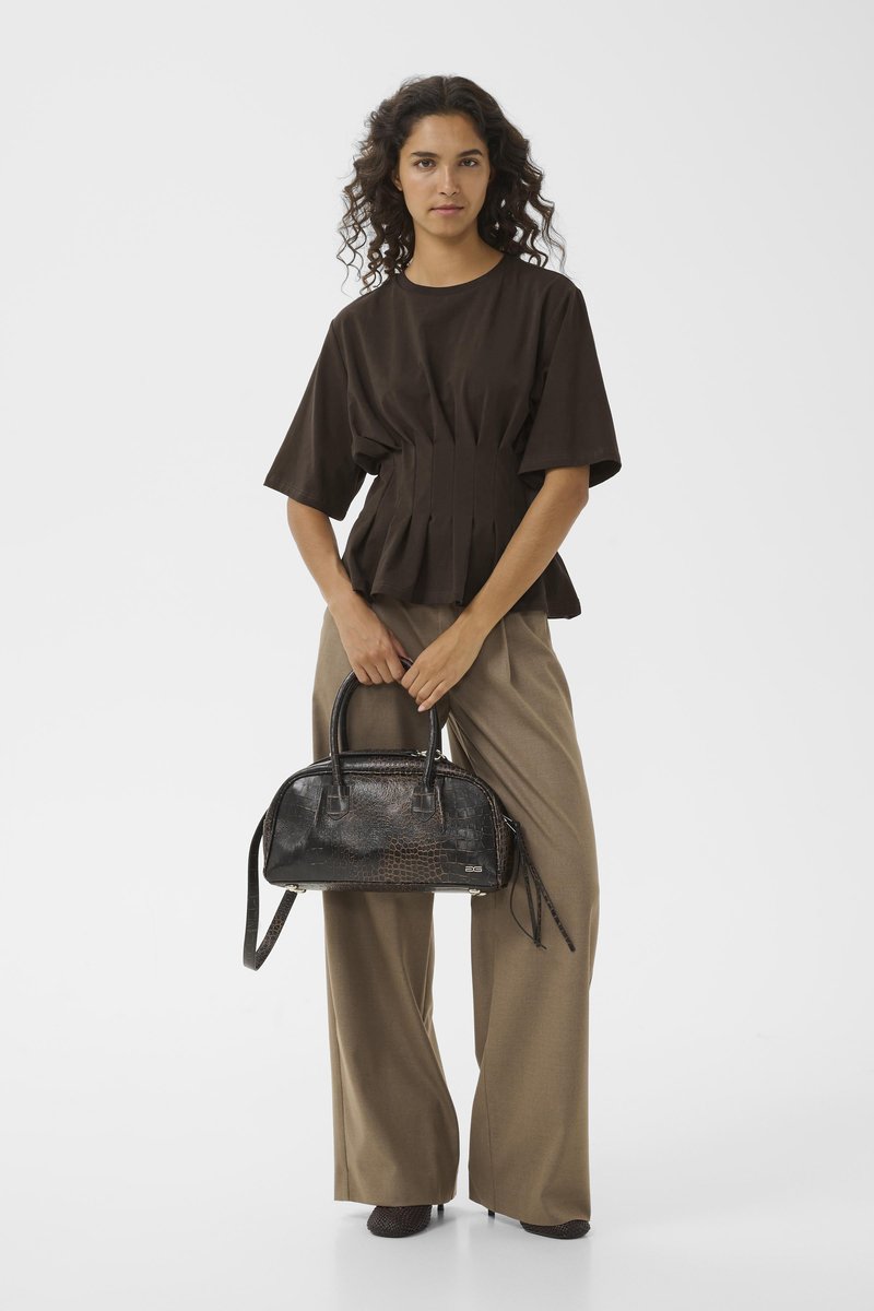 Haut plissé marron, pantalon large beige, tenant un sac à main en faux-crocodile noir avec des poignées courtes, surface texturée et finitions en métal argenté.