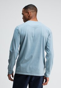 Helles blaues Langarmhemd aus Baumwollmaterial, mit Rundhalsausschnitt, taillierte Passform, sichtbare Nahtdetails, getragen mit dunklen Hosen, Rückansicht.