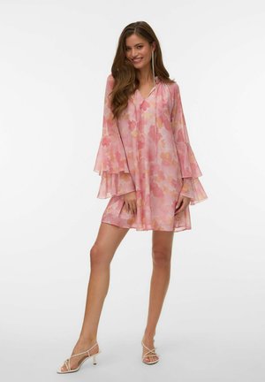 Femme portant une robe trapèze rose à fleurs avec des manches cloche superposées et des sandales blanches à talons avec lanières, souriant avec la main près de la cuisse.