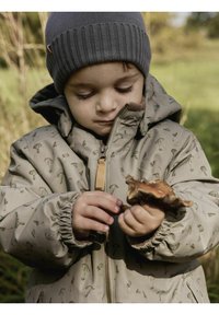 Beżowa kurtka w grzybowy wzór, z elastycznymi mankietami i zamkiem z przodu. Szara robiona na drutach czapka. Dziecko trzyma kawałek drewna, z głową lekko pochyloną w dół.